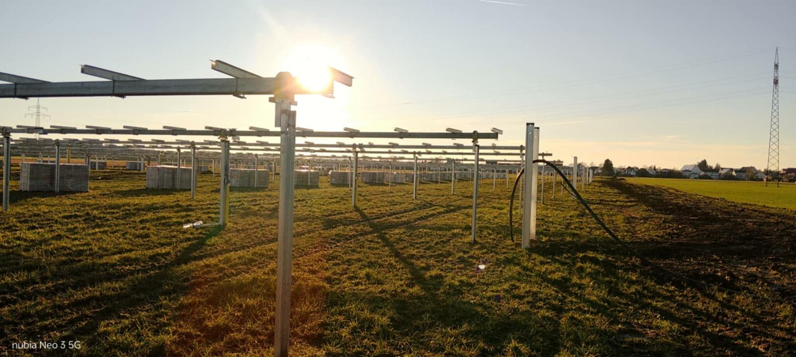 Im Bau befindliche Freiflächen-Photovoltaikanlage: Metallgestelle für die Solarmodule stehen in Reihen auf einer Wiese, im Hintergrund Materialkisten und Ortsrand, die tiefstehende Sonne spiegelt auf den Schienen.